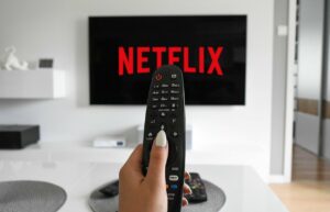 Imagem com foto de uma mão feminina segurando o controle remoto com o logo da Netflix ao fundo na televisão