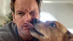 Imagem com foto do ator Patrick Wilson com um dos seus cachorros o lambendo