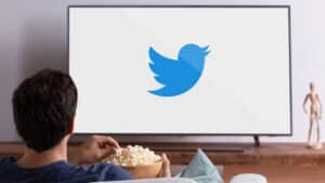 Imagem com foto de logotipo do Twitter na tela de um aparelho de televisão. O telespectador aparece sentado em frente o aparelho com um recipiente com pipoca