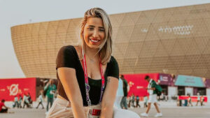 Imagem com foto da jornalista Isabelle Costa em frente a um dos estádios da Copa no Catar