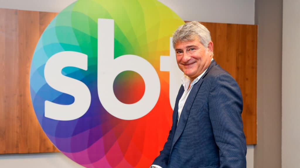 Cléber Machado em foto de divulgação com o logo do SBT ao fundo