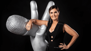 Fátima Bernardes com o braço direito apoiado no totem da mão que segura microfone do The Voice Brasil