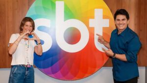 Foto de Daniela Beyruti e de Cesar Filho ao lado de um logo gigante do SBT