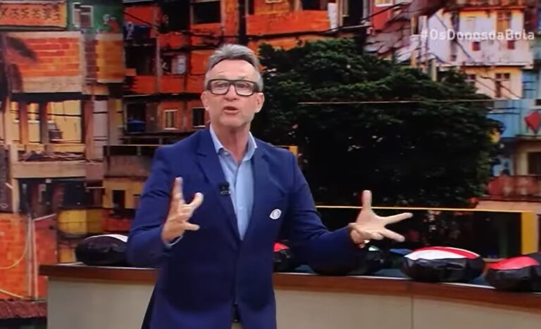 A imagem mostra o apresentador e ex-jogador Craque Neto em um cenário de estúdio, aparentemente em um momento de grande entusiasmo. Ele veste um blazer azul-escuro sobre uma camisa clara, gesticulando de forma expressiva enquanto fala