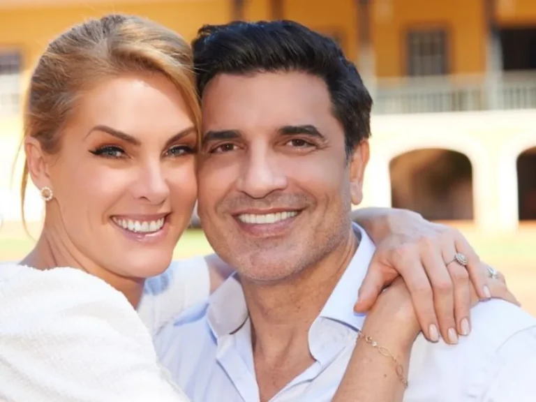 Na imagem, vemos Ana Hickmann e Edu Guedes em um momento feliz e descontraído. Eles estão sorrindo e abraçados, transmitindo uma sensação de cumplicidade e carinho. Ana Hickmann veste uma roupa clara e elegante, com acessórios delicados, incluindo brincos e pulseiras. Seu cabelo está preso, destacando seu rosto iluminado e expressivo