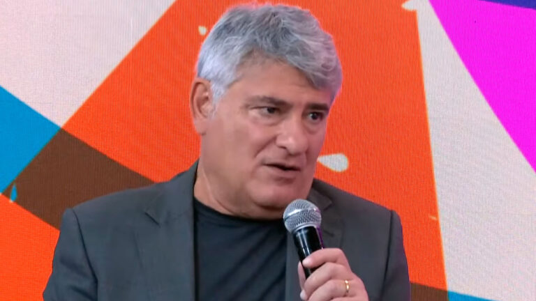 Cléber Machado, narrador da Record, participa do programa Pânico segurando microfone em frente a painel colorido