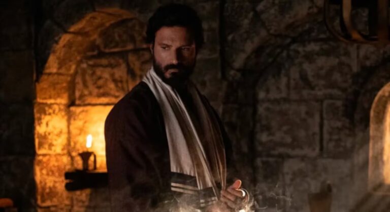 Na imagem, uma cena da série Paulo, o Apóstolo mostra o protagonista em um ambiente de iluminação baixa e atmosfera dramática. O personagem, interpretado por Murilo Cezar, aparece com expressão séria e olhar introspectivo, vestindo túnica escura e um manto claro sobre os ombros. Ao fundo, a parede de pedra e a luz de uma vela acesa reforçam o clima de época e a ambientação histórica da produção.