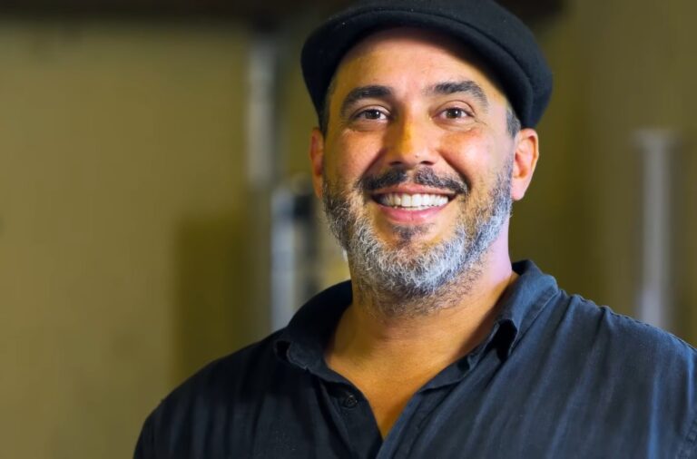 Na imagem, o apresentador André Marques aparece sorridente, usando uma camisa preta e um boné combinando. Ele está em um ambiente interno com iluminação suave, que destaca seu rosto iluminado e expressão descontraída.