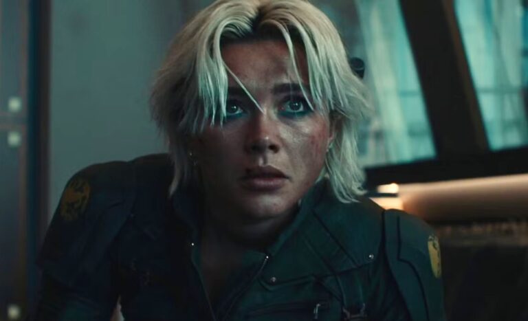 Florence Pugh aparece caracterizada como Yelena Belova em Thunderbolts. Com cabelos loiros curtos e maquiagem intensa nos olhos, a personagem surge em cena tensa, com o rosto sujo e marcas de batalha. Vestindo um uniforme tático escuro, ela transmite determinação e vulnerabilidade em meio a um ambiente de conflito.