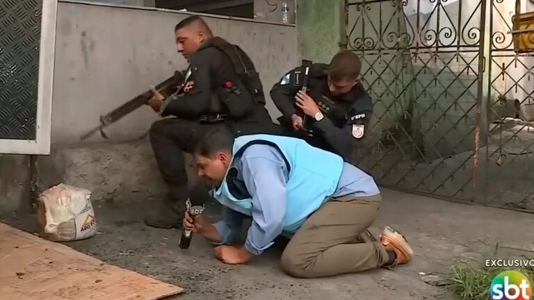 Jackson Silva com microfone do SBT abaixado ao lado de policiais armados durante cobertura no SBT Rio