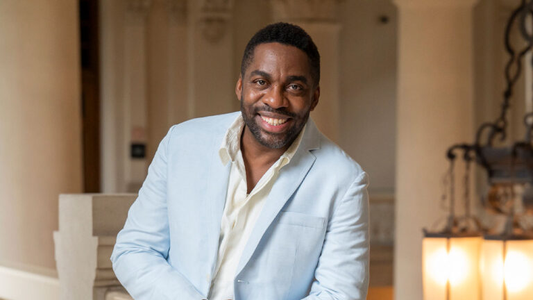 Lázaro Ramos sorrindo com blazer azul claro em ambiente interno iluminado com lustres ao fundo