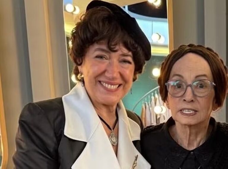 Lilia Cabral e Joana Fomm posam juntas em clima de celebração. Lilia sorri para a câmera, usando boina preta, brincos dourados e um elegante casaco preto com gola branca. Ao seu lado, Joana Fomm aparece de óculos de grau e cabelo preso, com expressão serena e carinhosa. O cenário de bastidores, iluminado por espelhos com luzes, sugere um encontro especial nos estúdios de gravação.