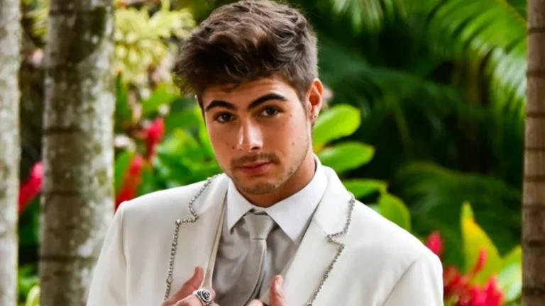 Rafa Vitti posa com elegância em cenário tropical, usando um terno branco com detalhes em brilho e gravata prata. O ator aparece em meio a plantas e flores, com expressão descontraída e olhar direto para a câmera.