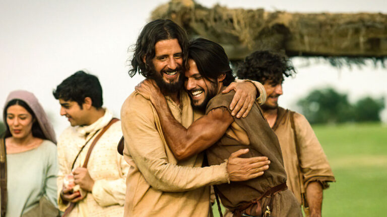 Cena da novela The Chosen com personagens usando trajes de época abraçados em campo aberto durante o dia