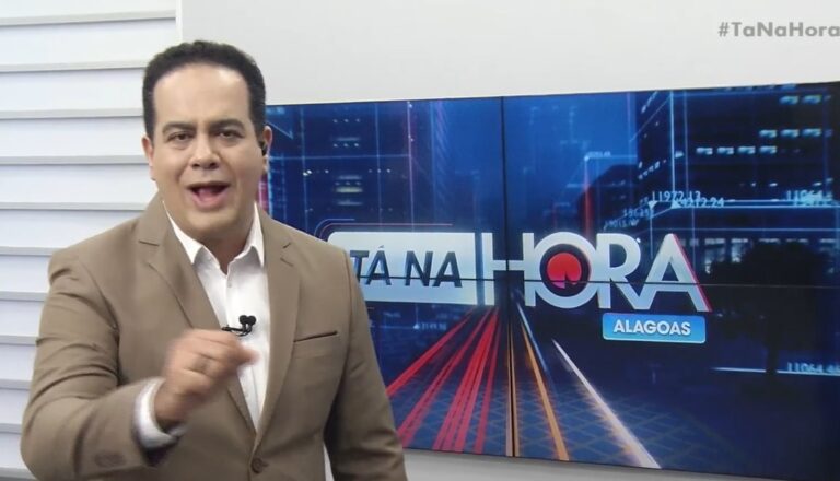 Wescley Costa, apresentador do Tá na Hora Alagoas, aparece em estúdio durante a exibição do telejornal. Vestido com terno bege e camisa branca, ele gesticula enquanto fala, com o logotipo do programa exibido em um telão ao fundo. A imagem transmite seu estilo comunicativo e enérgico, característico da atração exibida pela TV Ponta Verde, afiliada do SBT