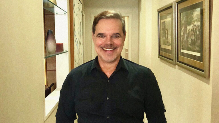 Diogo Vilela sorrindo em corredor decorado com quadros na parede e prateleira com vaso de cerâmica