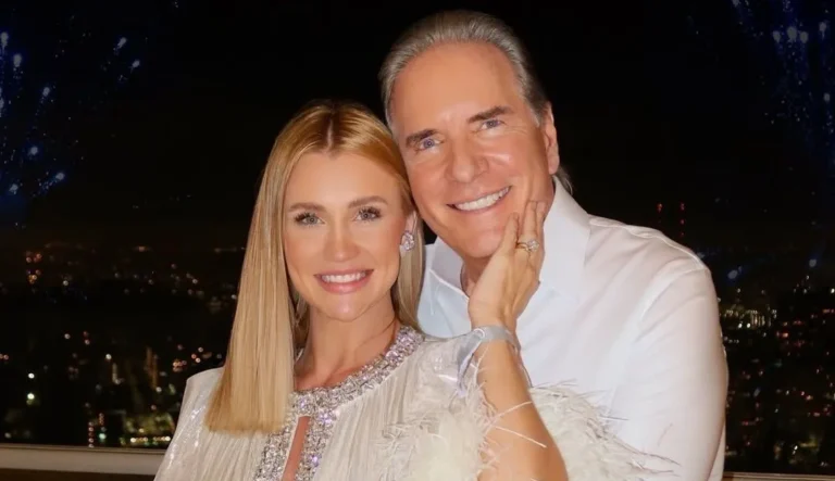 Na imagem, Roberto Justus e Ana Paula Siebert posam sorridentes e abraçados diante de uma vista noturna iluminada, com pontos de luz ao fundo que indicam a cidade e fogos de artifício no céu. Ana Paula usa um vestido branco com detalhes em brilho e plumas, enquanto Justus está com camisa social branca. Ambos exibem expressões de alegria e cumplicidade, reforçadas pela mão dela apoiada no rosto dele. O cenário e a produção visual sugerem um momento especial, possivelmente comemorativo.