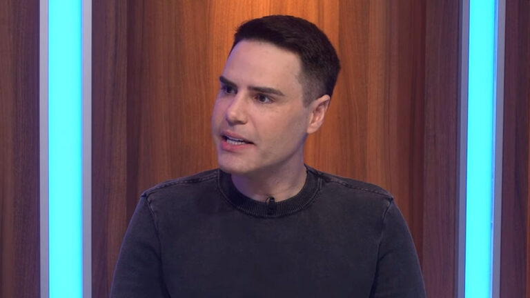 Luiz Bacci falando durante participação no Programa Flávio Ricco diante de painel com luzes azuis