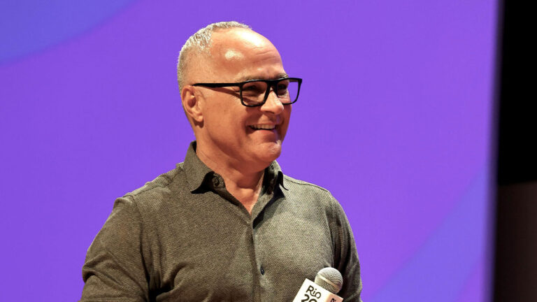 Amauri Soares sorri enquanto segura microfone durante evento com fundo roxo iluminado