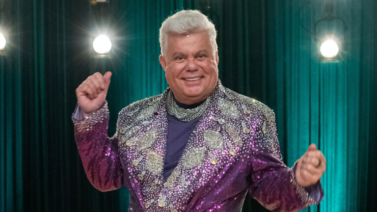 Milton Cunha sorrindo com paletó roxo brilhante em cenário com cortinas verdes e luzes de estúdio ao fundo