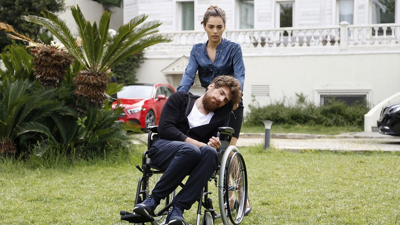 Nedim (Berker Güven) desacordado em cadeira de rodas sendo empurrado por Cemre (Sera Kutlubey) em Cidade Cruel