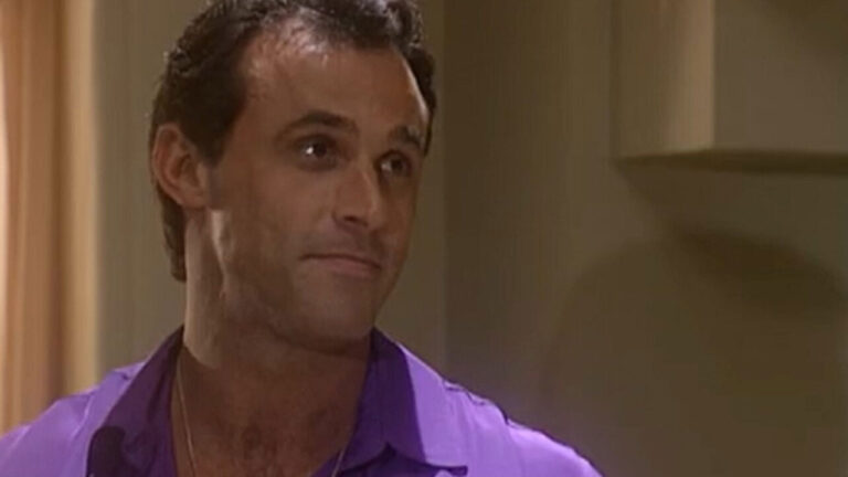 Oscar Magrini caracterizado como Ralf em cena de O Rei do Gado, usando camisa roxa e com expressão séria