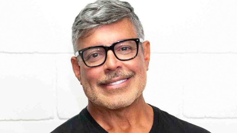 Alexandre Frota sorridente usa óculos de grau e camiseta preta posando em frente a parede branca