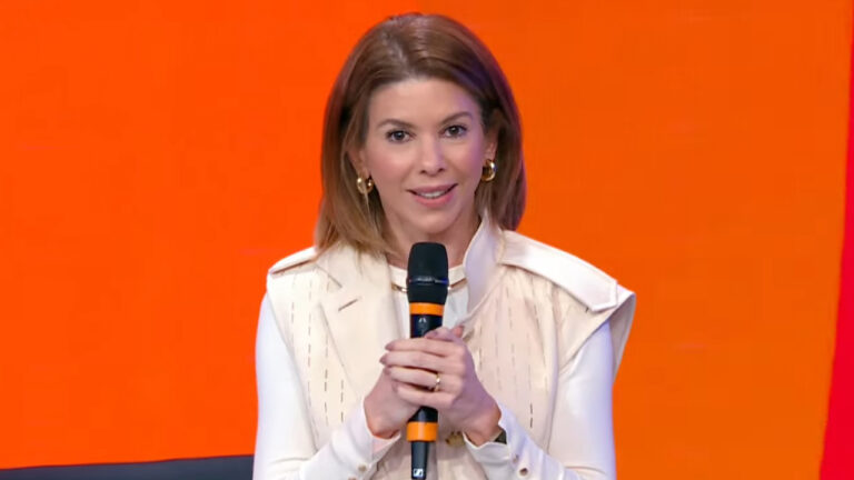 Daniela Beyruti, presidente do SBT, segura microfone e fala em evento com fundo laranja