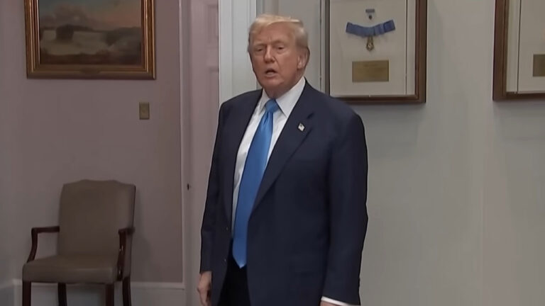 Donald Trump de terno escuro e gravata azul em pé dentro de sala com quadros e medalhas na parede