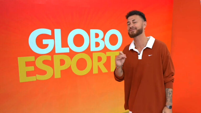 Fred Bruno em frente ao telão do Globo Esporte usando camisa polo larga marrom com detalhes brancos