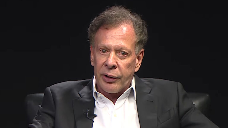 João Camargo de paletó escuro e camisa branca participa de entrevista em estúdio com fundo preto