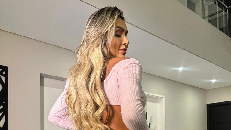 Mari Reis de cabelos loiros longos usando blusa rosa de mangas compridas em ambiente interno iluminado