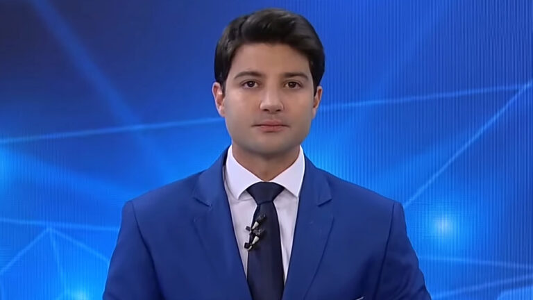 Rafael Algarte veste terno azul e gravata escura enquanto apresenta telejornal da Record News em estúdio com fundo azul geométrico