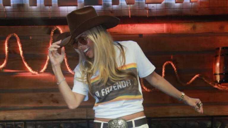 Adriane Galisteu com chapéu marrom e camiseta A Fazenda sorrindo no cenário do reality show da Record
