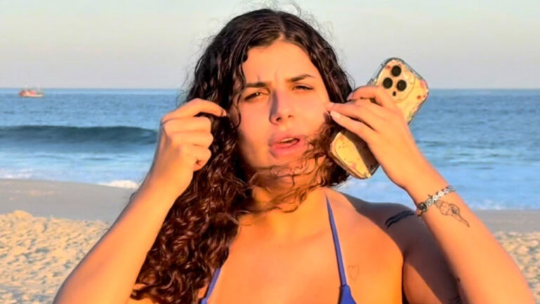Alê de biquíni azul segura celular no ouvido e ajeita o cabelo na praia com o mar ao fundo
