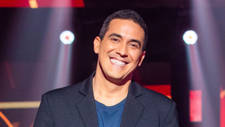 André Marques sorrindo de blazer preto e camiseta azul em estúdio com luzes coloridas ao fundo no cenário do The Voice+
