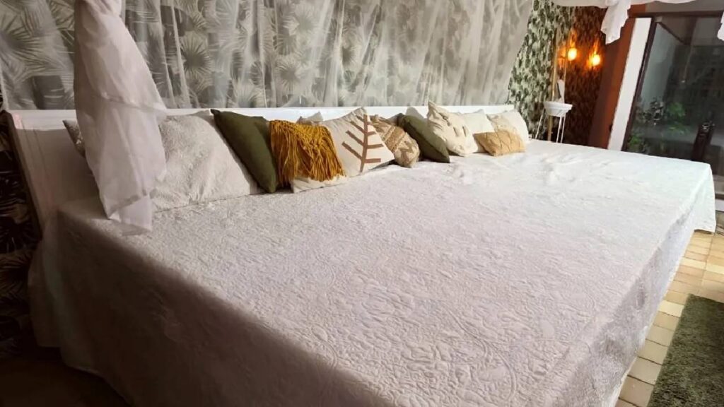Cama de 6 metros colocada à venda por Arthur O Urso, coberta por colcha branca e decorada com várias almofadas coloridas, em quarto da Mansão do Amor
