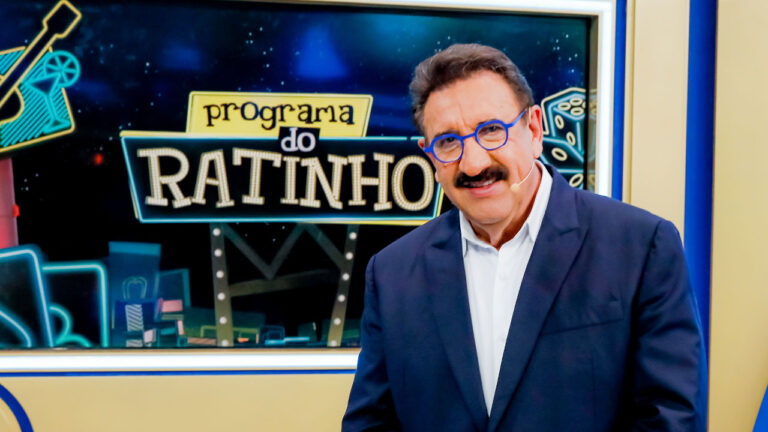 Carlos Massa, o Ratinho, de óculos azuis e terno escuro em frente ao letreiro do Programa do Ratinho no SBT