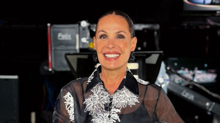 Carolina Ferraz sorrindo com blusa preta bordada em estúdio de televisão com equipamentos ao fundo