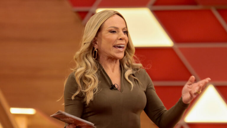 Christina Rocha com microfone de lapela e ficha nas mãos apresenta Casos de Família no SBT diante de cenário vermelho geométrico