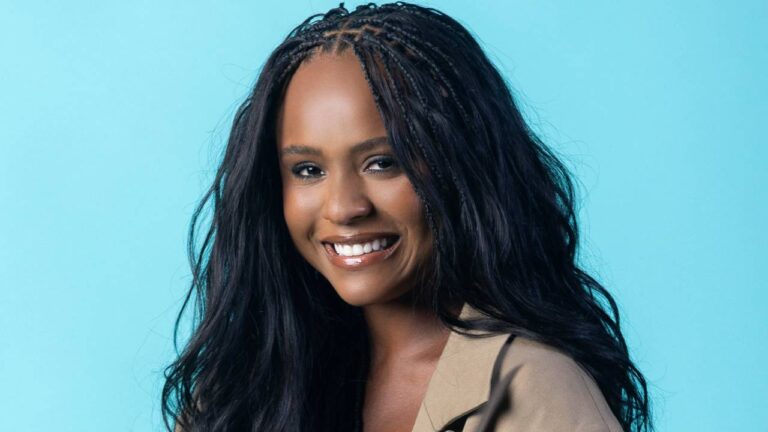 Duda Santos sorri em foto de estúdio com fundo azul usando casaco bege e cabelo solto com tranças na parte frontal