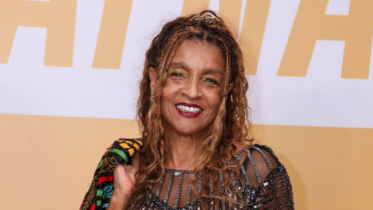 Elisa Lucinda sorrindo com vestido preto transparente bordado e detalhes brilhantes em evento com fundo amarelo