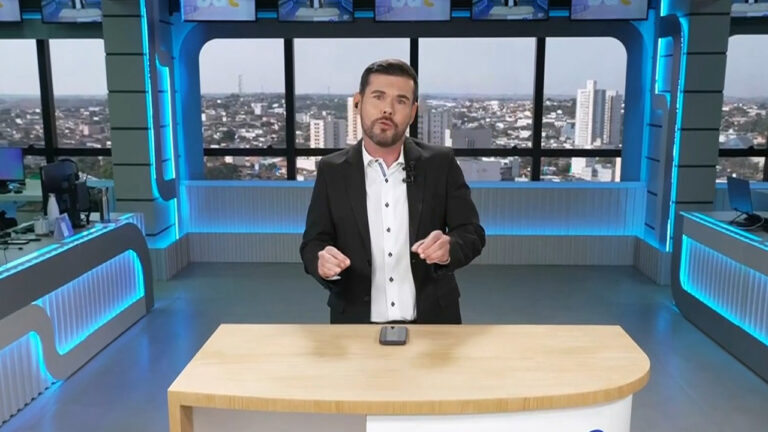 Emerson Sanchez de terno escuro apresenta o Bom Dia Cidade no estúdio da TV Tem em Presidente Prudente com janelas mostrando a cidade ao fundo