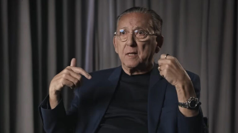 Galvão Bueno usando óculos e blazer azul escuro gesticulando durante entrevista em estúdio com fundo cinza