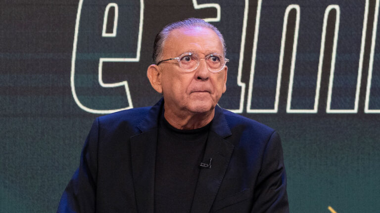 Galvão Bueno usa óculos transparentes e blazer preto em frente a painel com letras grandes iluminadas