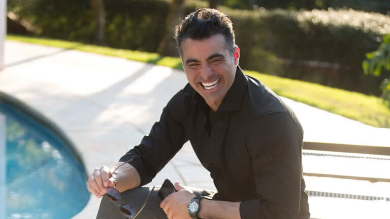 JP Mantovani sorrindo ao ar livre sentado perto de uma piscina segurando óculos escuros na mão direita