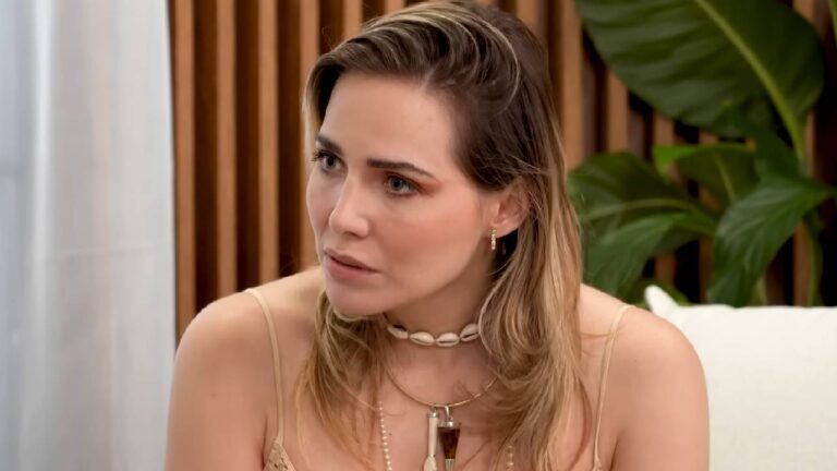 Letícia Colin com expressão séria usa colar de búzios e vestido claro sentada em ambiente interno com plantas ao fundo