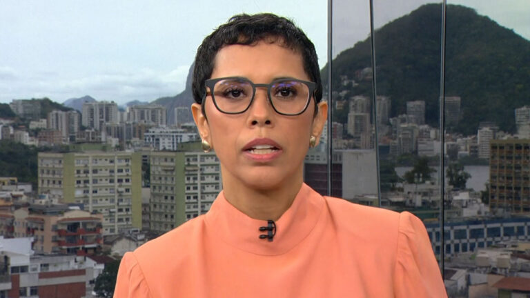 Lilian Ribeiro usa blusa salmão e óculos de grau enquanto apresenta o RJ1 com vista do Rio de Janeiro ao fundo