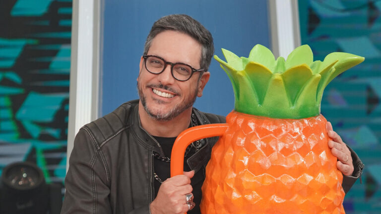 Lucio Mauro Filho sorridente de jaqueta preta segura caneca gigante em formato de abacaxi no estúdio do Caldeirão com Mion