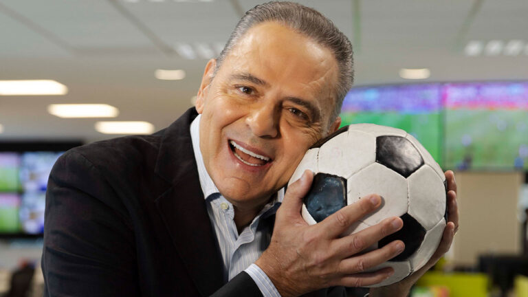 Luis Roberto sorrindo e segurando uma bola de futebol junto ao rosto em redação da Globo